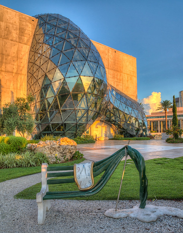 The geodesic glass Enigma dome of the Dalí Museum and a melting clock sculpture on a green park bench.
