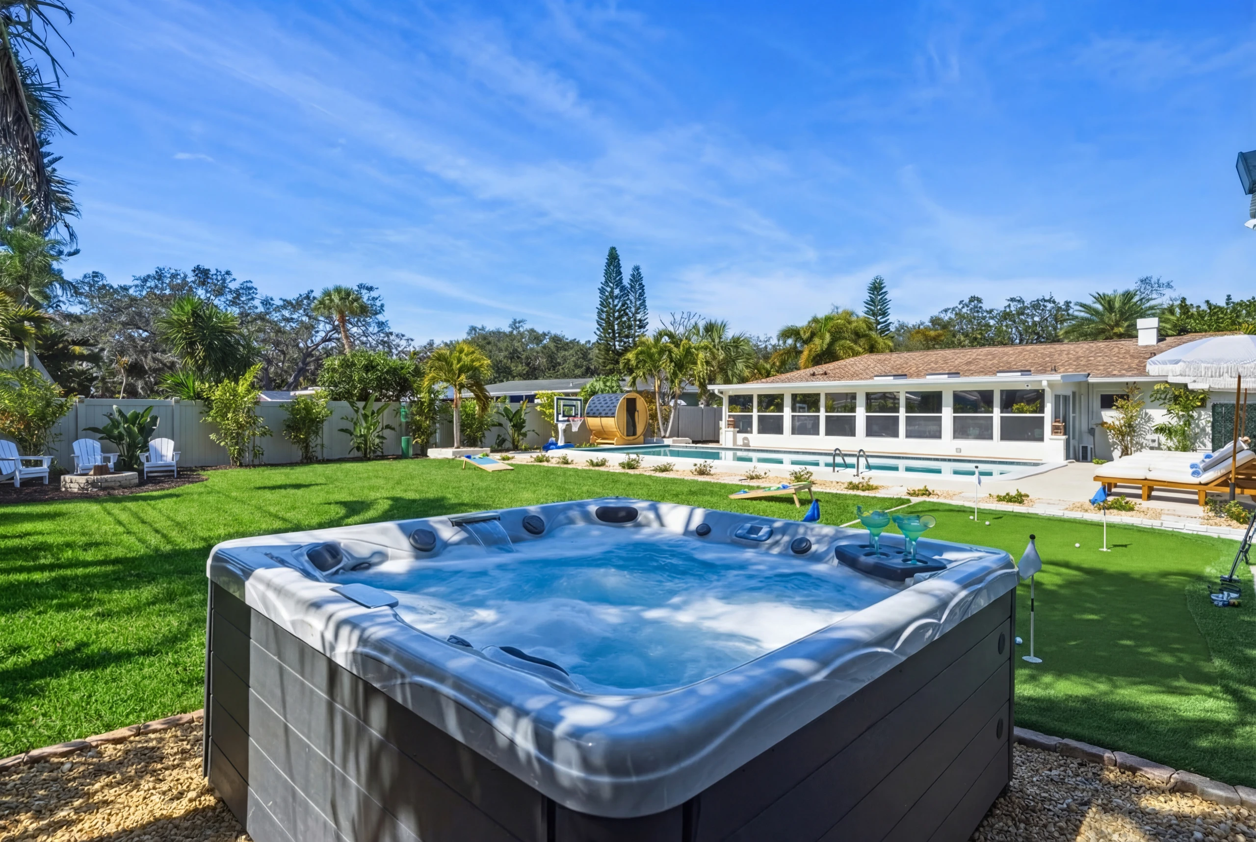 Illuminated outdoor hot tub at dusk with a putting green, swimming pool, and barrel sauna in a private backyard.