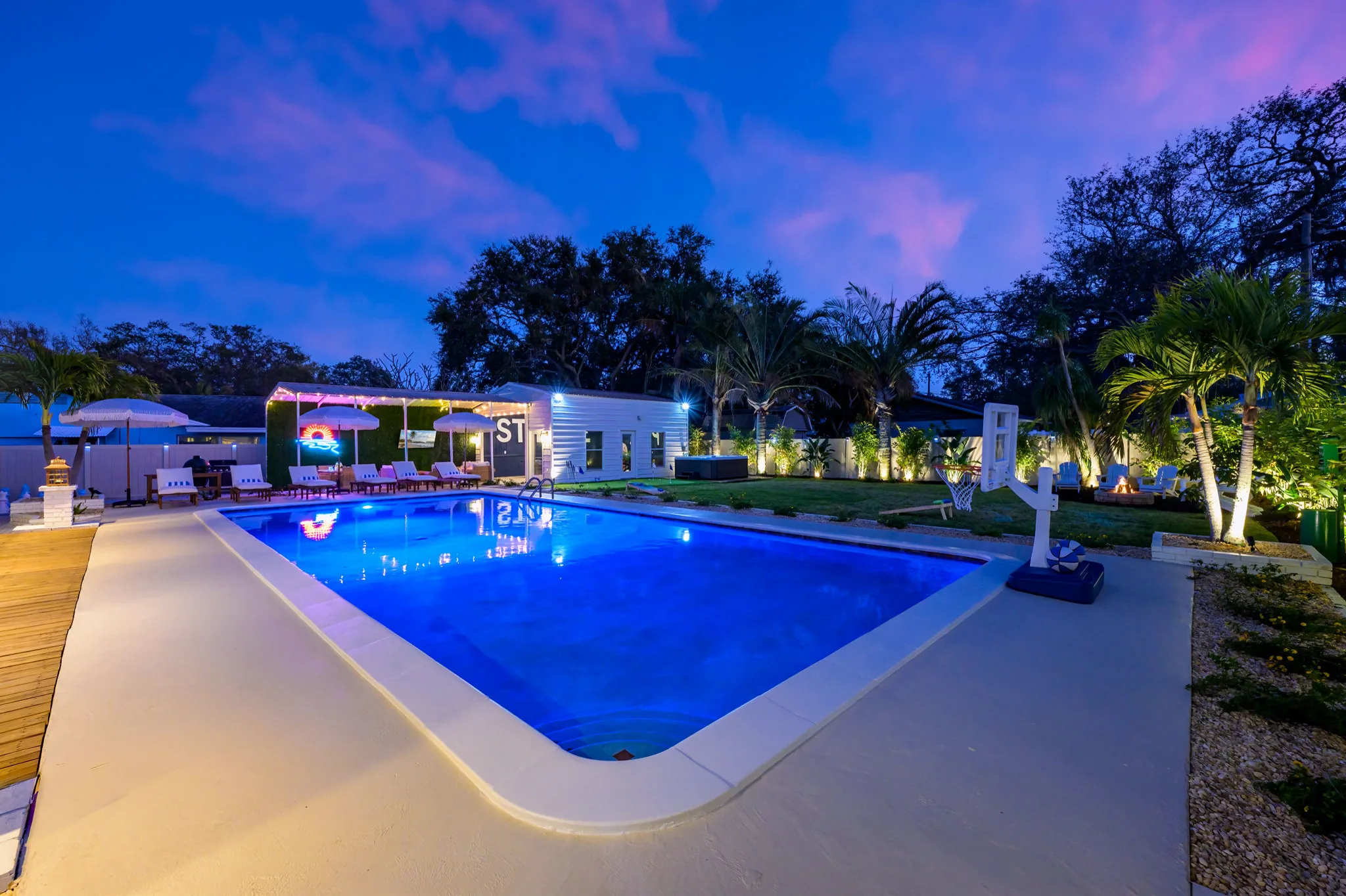 Large illuminated luxury swimming pool at dusk with poolside loungers, basketball hoop, and palm trees.